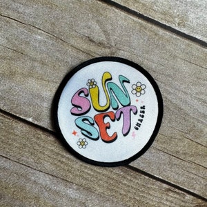 May include: A white round patch with black stitching around the edge. The patch has a colorful graphic of the word "SUNSET CHASER" in a retro font with flowers and stars.