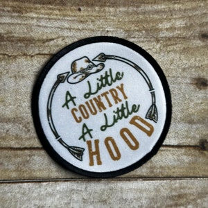 May include: A white round patch with black stitching around the edge. The patch has a brown and green camouflage pattern with a brown cowboy hat, two arrows, and the text "A little country a little hood" in gold lettering.