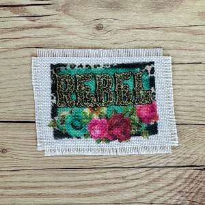 May include: A white canvas with a fringed edge featuring a colorful floral design and the word "Rebel" in black letters.