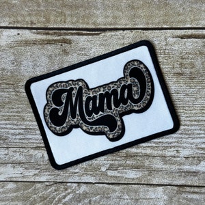 May include: White patch with black stitching, featuring the word "Mama" in a black, cursive font with a leopard print pattern.