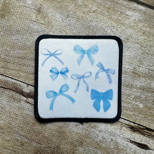 May include: A white patch with blue watercolor bows of various sizes and shapes. The patch is surrounded by a black border.