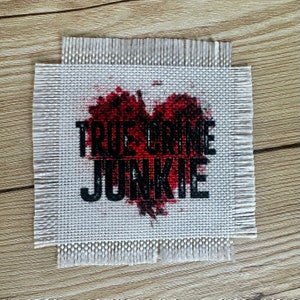 May include: A white fabric patch with a red heart-shaped splatter design and the text "TRUE CRIME JUNKIE" in black.