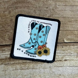 May include: A white patch with black edging features a pair of blue cowboy boots with white daisy patterns. The boots are surrounded by sunflowers and orange flowers. The text "In a world full of princesses, be a cowgirl" is printed on the patch.