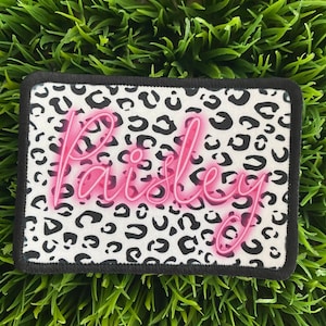 May include: A white and black leopard print patch with the name "Paisley" in pink neon lettering.