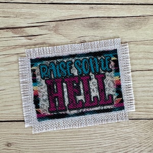 May include: A white fabric patch with a fringed border. The patch has a colorful geometric pattern and the text "Raise Some Hell" in blue, pink, and black.