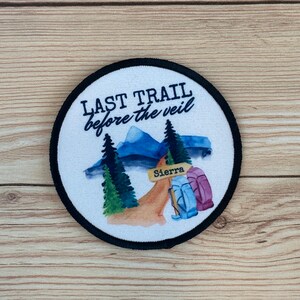 May include: A white round patch with black stitching around the edge. The patch features a watercolor illustration of a mountain path with two backpacks, trees, and a sign that reads "Sierra". The text "Last Trail before the veil" is written in black above the illustration.