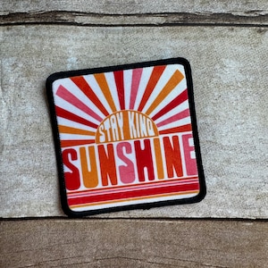 May include: A red, orange, and pink sunburst design with the text "Stay Kind Sunshine" on a black and white patch.