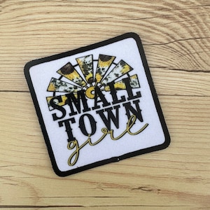 May include: A white square patch with black stitching around the edges. The patch features a black and white sunflower design with a yellow center. The text "SMALL TOWN girl" is written in black and yellow lettering.