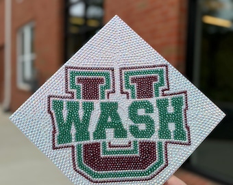 May include: A square graduation cap covered in clear rhinestones. The cap features the word "WASH" in green and maroon, with a green outline. The background is blurred, showing a brick building and windows.