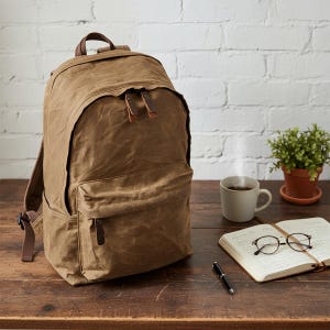 May include: A brown waxed canvas backpack with leather accents. The backpack has a large main compartment, a front pocket, and adjustable straps. A cup of coffee, a notebook, and glasses sit on a wooden table.