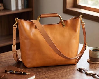 May include: A tan leather tote bag with a shoulder strap and brass-colored hardware. The bag has a rectangular shape with a top handle and a long, adjustable strap. A notebook and pen are on the wooden surface.