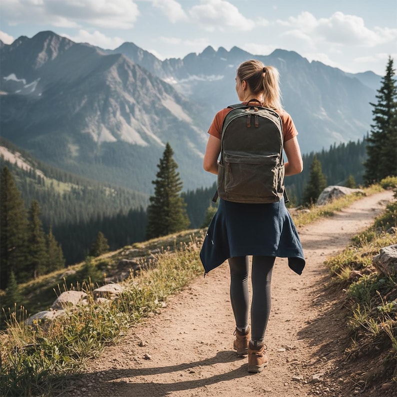May include: A person wearing a backpack walks on a dirt path in the mountains. The backpack is olive green. The person is wearing an orange shirt, dark leggings, and brown boots. The background features a mountain range and trees.