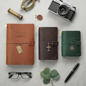 Könnte beinhalten: Drei Leder-Notizbücher in Hellbraun, Dunkelbraun und Grün mit Prägungen und Metallanhängern. Dazu eine Pentax-Kamera, ein Messingrohr, eine Brille und ein schwarzer Stift. Das hellbraune Notizbuch trägt das Wort Adventure, das dunkelbraune einen Kompass und das grüne Welcome Adventure.