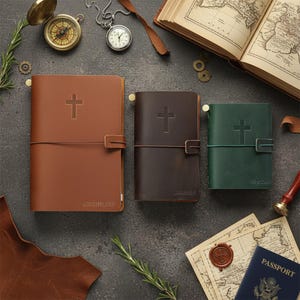 Peut inclure: Trois carnets reliés en cuir de couleur fauve, brun foncé et vert, chacun orné d'une croix en relief sur la couverture. Le carnet fauve porte l'inscription ADVENTURE, le brun JOURNEY et le vert EXPLORE. Ils possèdent des fermetures élastiques.