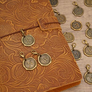 May include: A tan leather journal with an embossed floral pattern and a brown elastic strap, accompanied by several bronze-colored metal charms shaped like wax seals, each featuring a different embossed letter of the alphabet and a lobster clasp.