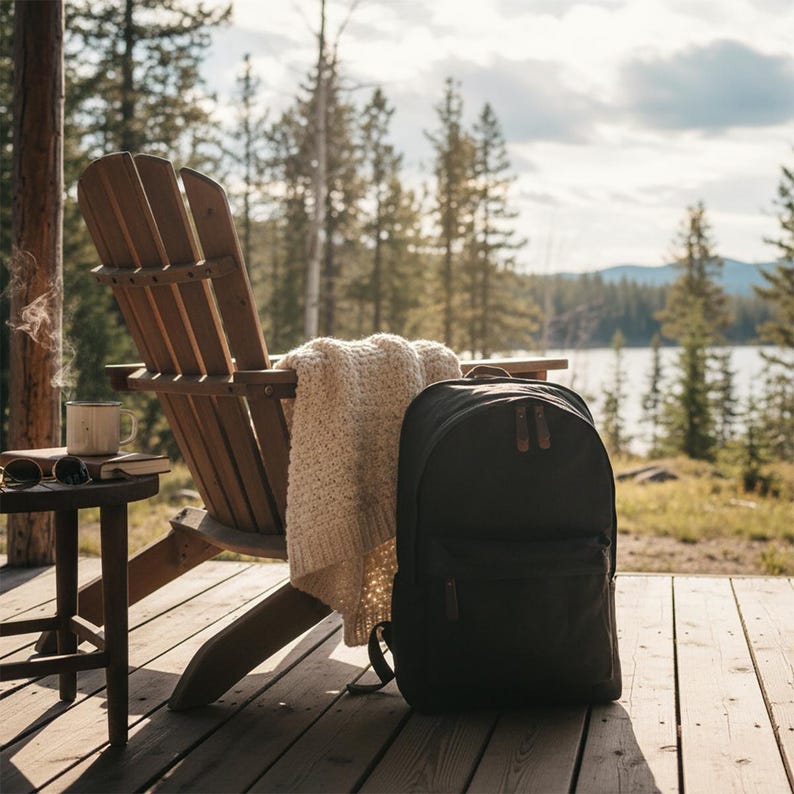 May include: A black backpack with brown leather accents sits on a wooden deck next to a wooden Adirondack chair with a cream-colored blanket. A small side table holds a mug, book, and sunglasses. The scene is set outdoors with a lake and trees in the background.