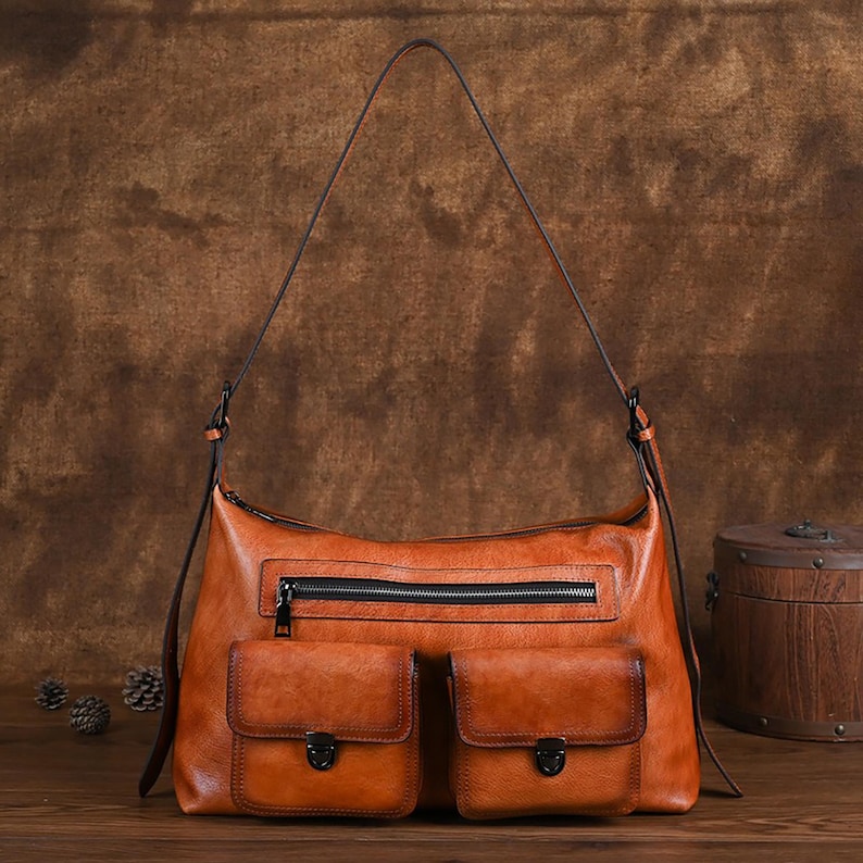 Op de afbeelding: Een bruine leren schoudertas met twee voorzakken en een ritssluiting. De tas heeft een verweerde look en een lange schouderband.