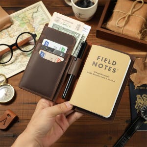 May include: A brown leather wallet with a Field Notes memo book, pen, and cards. The wallet is open, revealing cash and a Route 66 Adventure ticket. The memo book reads "FIELD NOTES" and "Durable Materials / Made in the U.S.A."
