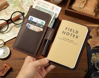 May include: A brown leather wallet with a Field Notes memo book, pen, and cards. The wallet is open, revealing cash and a Route 66 Adventure ticket. The memo book reads "FIELD NOTES" and "Durable Materials / Made in the U.S.A."