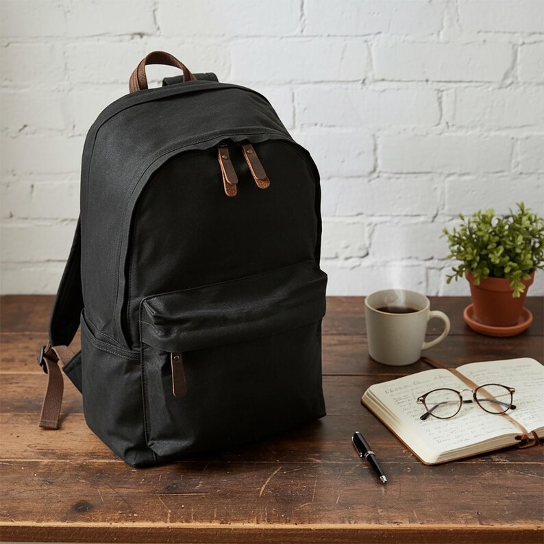 May include: A black backpack with brown leather accents, including the top handle and zipper pulls. The backpack is sitting on a wooden surface next to a cup of coffee, a notebook, and a pen.