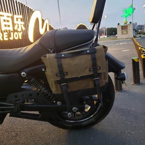 May include: A tan canvas motorcycle saddlebag with black leather straps and trim, mounted on the side of a black motorcycle. The bag features a rectangular flap secured by two vertical leather straps with metal buckles and rivets.