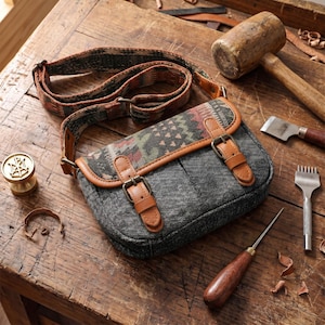 Pode incluir: Uma bolsa transversal cinza com aba estampada e detalhes em couro marrom. A bolsa tem uma alça estampada e dois fechos de fivela. Está sobre uma superfície de madeira com ferramentas de couro.
