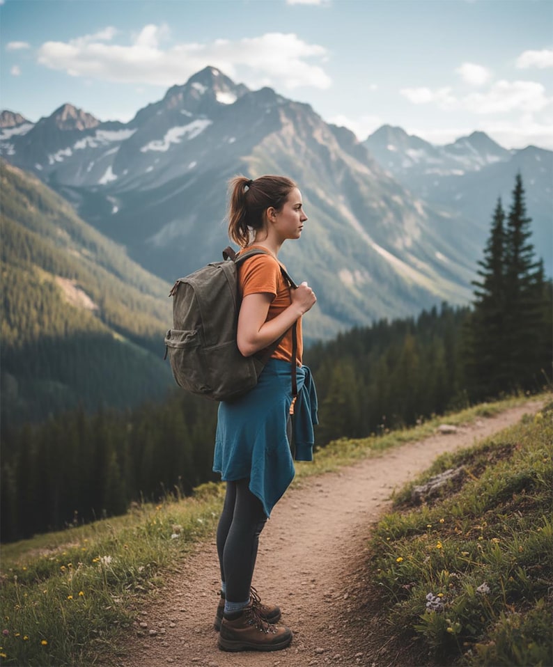 May include: A woman wearing an orange shirt, blue sweater, and black leggings stands on a dirt path, with a backpack. Brown hiking boots are visible. The background features a mountain range with snow-capped peaks and a forest.