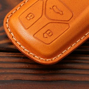 May include: A close-up of a brown leather key fob with stitching around the edges. The fob has three buttons, each with a raised design.