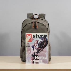 May include: Olive green canvas backpack with brown leather accents and straps. The backpack has a large main compartment and multiple pockets. A magazine with the word "stern" is propped up in front of the backpack.
