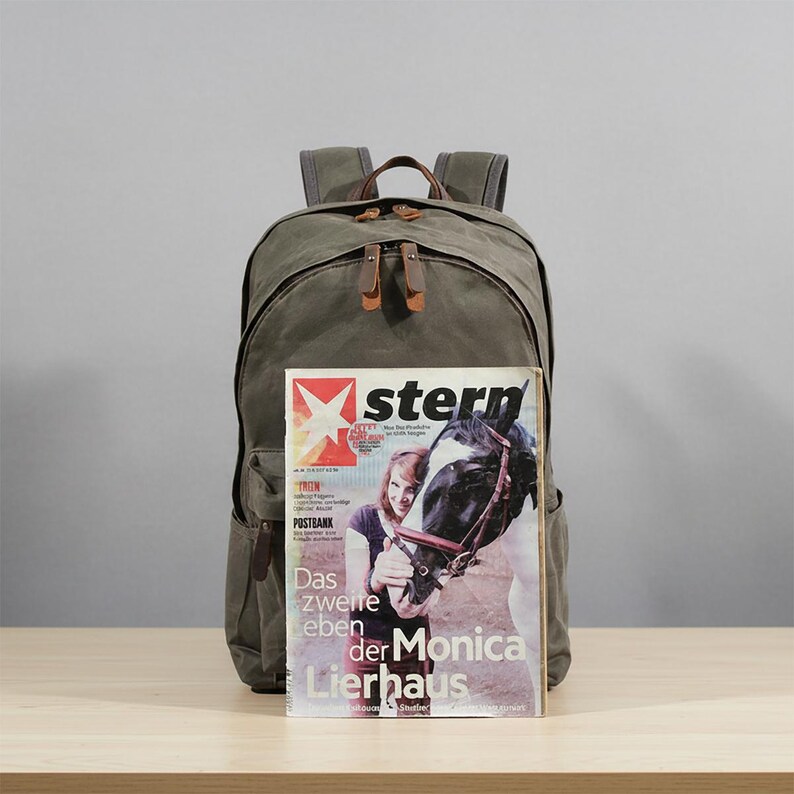 May include: Olive green canvas backpack with brown leather accents and straps. The backpack has a large main compartment and multiple pockets. A magazine with the word "stern" is propped up in front of the backpack.
