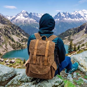 May include: A brown canvas backpack with tan leather straps, a top flap with buckle closures, a drawstring main compartment, and two side pockets. The backpack features a structured rectangular shape and is worn on the back with adjustable shoulder straps.