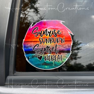 May include: A round sticker with a sunset scene, featuring the text "Sunrise, Sunburn, Sunset, Repeat" in black script. The background shows a vibrant sunset over the ocean, with shades of orange, pink, and blue. A heart symbol is included.
