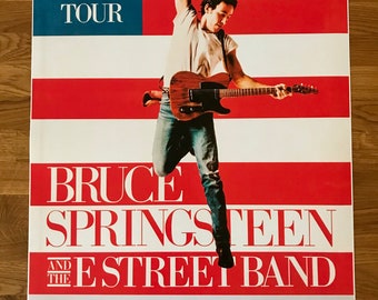 May include: A vintage concert poster for Bruce Springsteen and the E Street Band's 1985 Born in the U.S.A. tour. The poster features a red, white and blue American flag background with a photo of Bruce Springsteen jumping in the air while playing a guitar. The poster lists tour dates and locations in Europe.