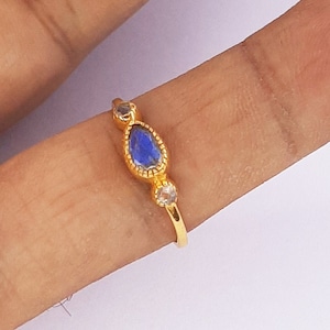 May include: A gold ring featuring a teardrop-shaped blue gemstone, framed by a gold border. Two smaller, clear gemstones flank the blue stone. The ring is displayed on a finger against a white background.