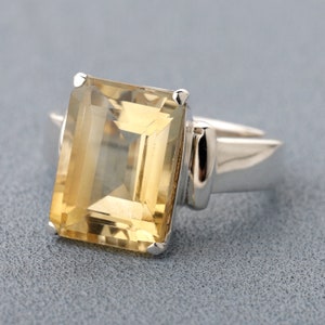 May include: A silver ring with a large, clear, yellow gemstone. The gemstone is cut in an emerald shape.