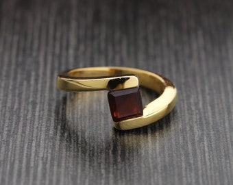 May include: A gold ring featuring a rectangular, dark red gemstone. The ring has a contemporary design, with the gemstone set in a bezel. The band curves around the finger, creating a unique and elegant appearance. The ring is a piece of jewellery.