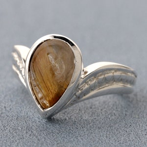 May include: A silver ring featuring a pear-shaped, golden-brown gemstone. The ring has a unique, curved design with decorative detailing on the band. The gemstone has a network of fine, golden inclusions.