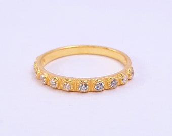 May include: A gold-colored ring with a series of small, clear gemstones set along the band. The ring has a textured design between each gemstone, adding detail to the piece. The ring is displayed on a white background.