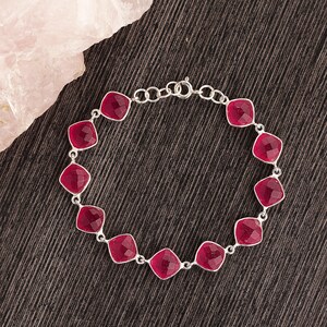 May include: A silver bracelet featuring square, faceted, ruby-red gemstones. The gemstones are set in a silver chain, creating a delicate and elegant piece of jewelry. The bracelet is displayed on a dark, textured surface.
