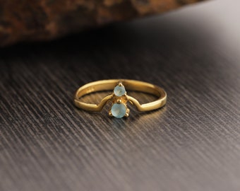 May include: A gold-colored ring with a unique design. The ring features two light blue gemstones, one larger than the other, set in a decorative setting. The ring is on a dark wood-grain surface.