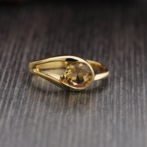 Op de afbeelding: Een gouden ring met een ronde, lichtbruine edelsteen. De ring heeft een uniek ontwerp met een gebogen band die om de steen heen loopt. De ring is geplaatst tegen een donkere, getextureerde achtergrond.