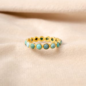 以下が含まれることがあります： 金色のリングで、ターコイズ色の石が並んでいます。リングのデザインは、ターコイズの宝石が連続して並んでおり、それぞれが互いに近くに配置されています。ターコイズの石には色のわずかな違いがあります。リングは柔らかく、ニュートラルな色の表面に置かれています。