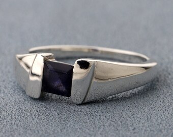 Può includere: Un anello color argento lucido con una singola pietra preziosa viola scuro a taglio quadrato, incastonata tra due griffe metalliche lisce e affusolate. La fascia presenta una struttura solida ed elegante con una finitura riflettente che mette in risalto la pietra.
