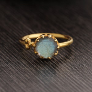 Può includere: Un anello d'oro con una pietra preziosa di labradorite blu incastonata in una lunetta a forma di conchiglia. L'anello ha un piccolo gruppo di perle dorate sul lato.