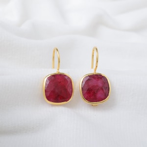 Puede incluir: Un par de pendientes colgantes dorados con piedras preciosas cuadradas, facetadas, de color rojo intenso. Los pendientes están engastados en un bisel dorado y cuelgan de un sencillo gancho dorado. El fondo es una tela blanca suave.