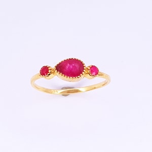 Puede incluir: Un anillo de oro con una gema grande en forma de lágrima de color magenta, flanqueada por dos gemas redondas más pequeñas de color magenta. El anillo tiene una banda fina y un engaste biselado texturizado.