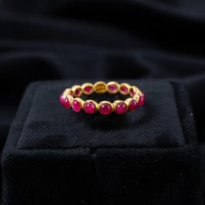 Könnte beinhalten: Ein goldener Ring mit einer durchgehenden Reihe leuchtend roter Edelsteine. Der Ring liegt auf einer schwarzen Samtoberfläche, die das Design und den Farbkontrast des Schmuckstücks hervorhebt. Die Edelsteine sind gleichmäßig angeordnet.