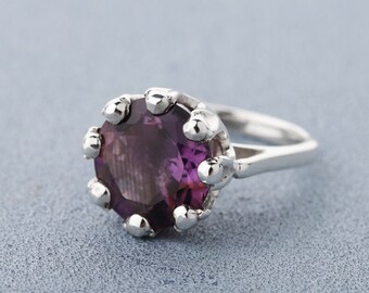 May include: A silver ring featuring a large, round, deep purple gemstone. The gemstone is set in a crown-like setting with small, rounded silver prongs. The ring band is smooth and polished, set against a light blue background.