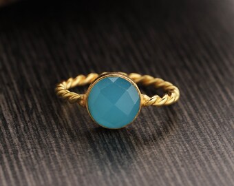 May include: A gold-colored ring with a round, faceted, light blue gemstone. The band has a twisted rope design. The ring is set against a dark wood grain background.
