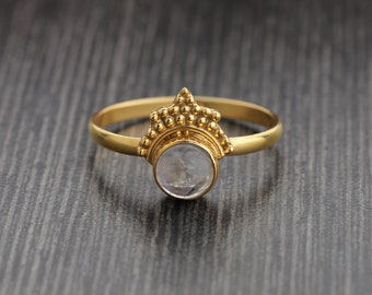 Puede incluir: Un anillo dorado con una piedra redonda y clara. El anillo tiene un diseño decorativo en forma de corona sobre la piedra. El fondo es de color gris oscuro.